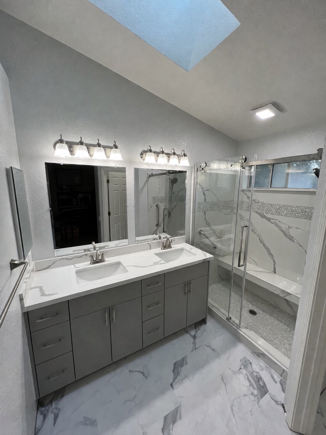 Primary bathroom after renovation with gray double vanity, stone surfaces, and glass shower